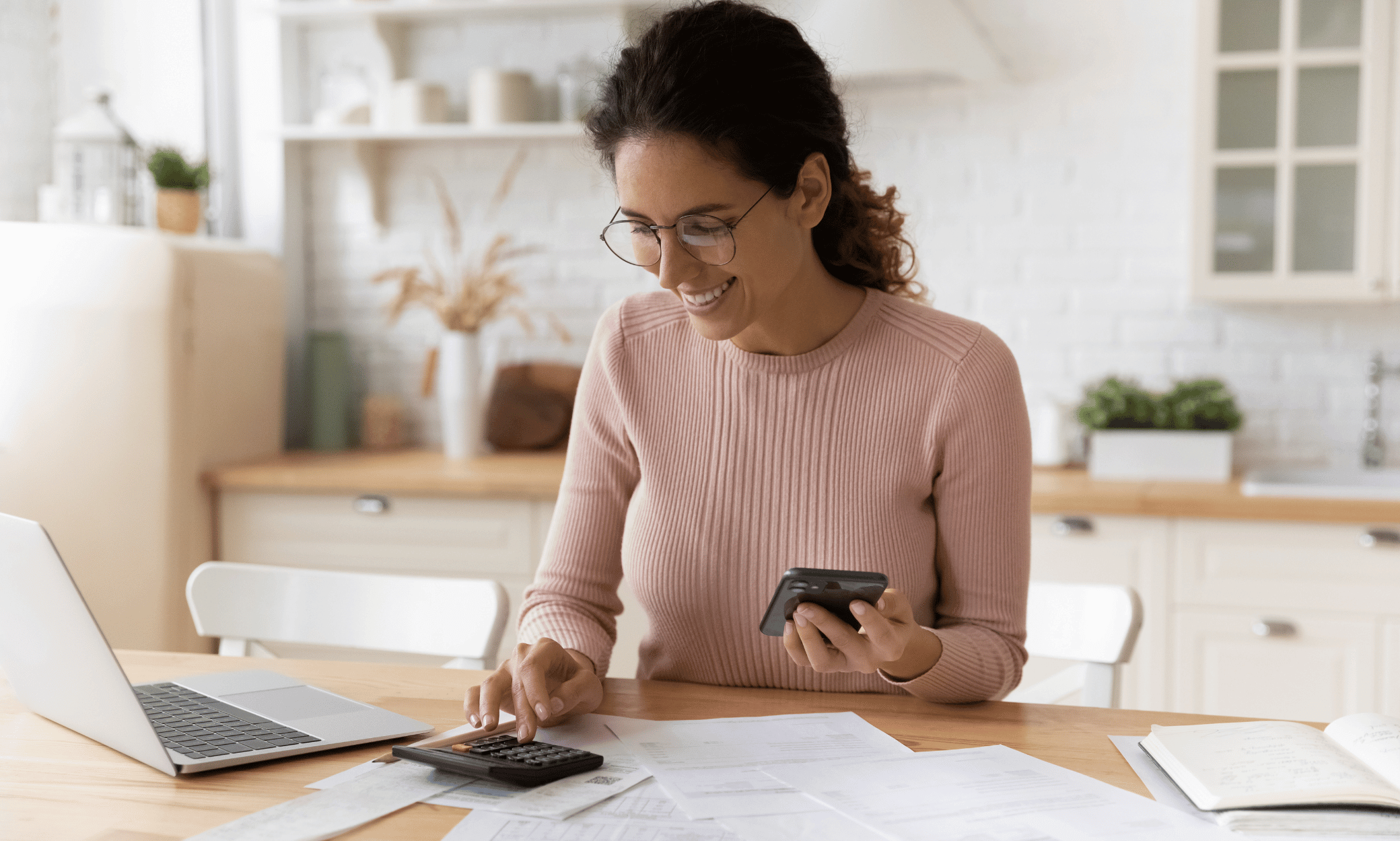 A person calculating work with her calculator, phone, and laptop while smiling in a pink top and glasses.